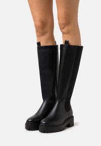 Schwarze kniehohe Stiefel aus glattem Leder mit robusten Sohlen, ausgestattet mit Ziehschlaufen und einem runden Zehendesign. Strukturiertes Finish an der Außenseite.