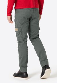Grüne Cargo-Hose mit schlanker Passform, seitlichen Taschen und einem Patch mit Branding. Kombiniert mit schwarzen Schuhen und einem roten Langarmshirt.