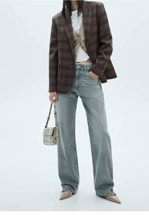 Femme portant un blazer à carreaux marron, un cardigan beige, un haut blanc, un jean gris ample, des chaussures beige à bouts pointus, tenant un petit sac à main en peau de serpent.