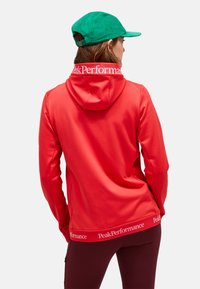 Rode hoodie met een capuchon en een wit merklabel aan de kraag. Draagt met een groene pet; de stof lijkt glad, heeft een strakke pasvorm en bevat logo-accenten.