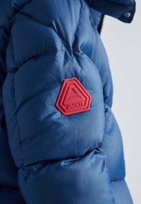 Pufferjack met een glanzende blauwe afwerking, voorzien van gestikte secties en een rode hexagonale patch met de tekst "JACKIT" op de mouw.