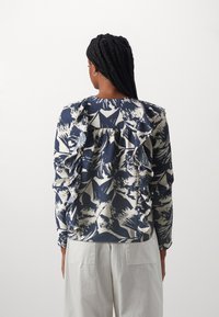 Maglia a maniche lunghe blu navy e crema con motivo floreale, ruches testurizzate sulla parte posteriore e scollatura rotonda. Pantaloni bianchi visibili nella parte inferiore.