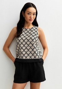 Top negro sin mangas con patrón floral blanco, escote redondo y dobladillo negro en contraste. Combinado con pantalones cortos negros, la tela parece texturizada.