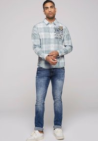 Hellblau kariertes Hemd mit langen Ärmeln und Button-Down-Kragen. Hat einen schwarzen grafischen Druck auf der Brust. Wird mit faded blauen Jeans und Sneakers getragen.