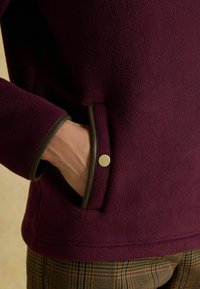 Roka atpūta iekšējos sānu kabatā bordo vilnas mēteļa ar brūnu apdari un zelta pogu, zemāk redzamas svītrainas bikses.