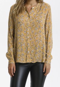 Senfgelbe Bluse mit Knöpfen und einem floralen Muster in Weiß und Schwarz. Sie hat einen runden Kragen und eine plissierte Textur. Lange Ärmel.