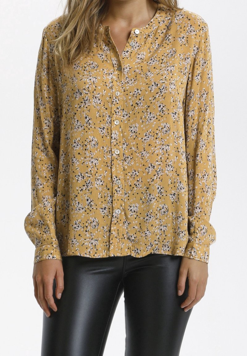 Senfgelbe Bluse mit Knöpfen und einem floralen Muster in Weiß und Schwarz. Sie hat einen runden Kragen und eine plissierte Textur. Lange Ärmel.