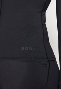 Μαύρο αθλητικό τοπ με ριγωτή υφή και λεπτομέρεια λογότυπου. Απαλή υφή με εφαρμοστό σχεδιασμό, κατάλληλο για αθλητική ένδυση.