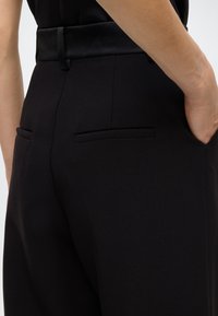 Črne hlače po meri z gladko teksturo, s pasom iz satena in zadnjimi žepi, ki zagotavljajo strukturiran kroj in eleganten dizajn.