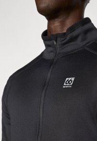 Zwarte fleece jas met een hoge kraag en ritssluiting. Beschikt over een klein wit logo op de borst en een gladde, gestructureerde stof.