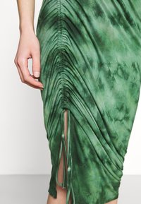 Main à côté d'une jupe verte froncée, teinte par nouage, avec une fente latérale et des cordons ajustables, montrant la texture du tissu et les fronces.