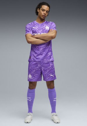 Lila Fußballtrikot mit einem wirbelnden Muster, bestehend aus einem kurzärmeligen Shirt und Shorts, kombiniert mit knielangen Socken und Fußballschuhen.