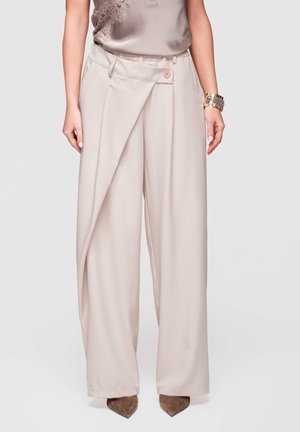 Donna che indossa pantaloni beige a vita alta e gambe larghe con dettaglio a pattina frontale e bottone, abbinati a scarpe marroni con punta affusolata.