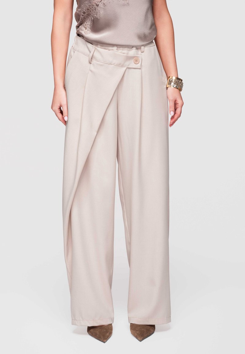 Femme portant un pantalon beige taille haute à jambes larges avec un détail de chevauchement à l'avant et un bouton, associé à des chaussures marron pointues.