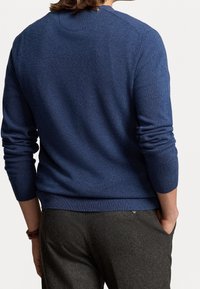 Personne portant un pull bleu à manches longues et un pantalon gris foncé, debout avec une main dans la poche, vue de dos.
