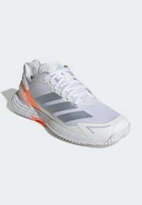 Valkoinen urheilukenkä, jossa on verkkomateriaalia yläosassa, harmaat kolme raitaa, oranssi yksityiskohta pohjassa ja teksturoitu kumipohja. Kevyt muotoilu.