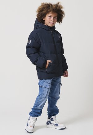Marineblauwe pufferjas met een capuchon, voorzien van een ritssluiting aan de voorkant en twee zakken. gedragen met lichtblauwe cargobroeken en witte sneakers met marineblauwe accenten.