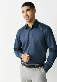 Mężczyzna uśmiechający się, ubrany w granatową koszulę z długim rękawem, włożoną w jasnoszare spodnie z czarnym paskiem, stojący na białym tle.