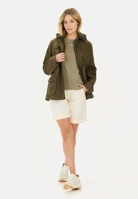 Grøn vandtæt jakke med hætte, iført åben over en olivengrøn t-shirt og cremefarvede shorts. Modellen har beige sneakers på.