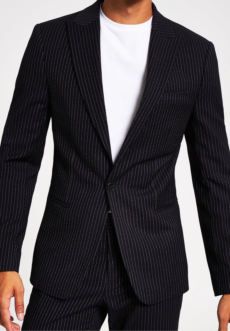 Mörkblå blazer med smårandigt mönster, enstaka knappstängning, slagkrage och bröstficka. Bärs över en vit t-shirt med rund halsringning.