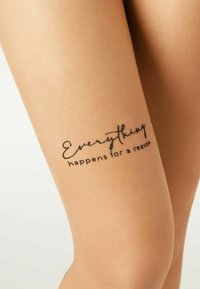 Beiga strumpbyxor med en slät textur som har ett handskrivet citat i svart: "Allt händer av en anledning," placerat på lårområdet.