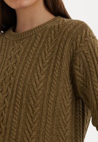 Καφέ πλεξούδα πουλόβερ με καλωδίωση, στρογγυλή λαιμόκοψη και ριμπ μανσέτες. Η υφή είναι απαλή και χοντρή, κατάλληλη για δροσερό καιρό.