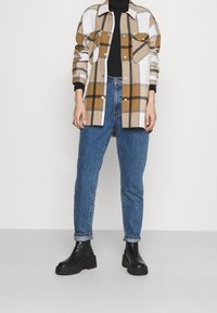 Camicia a quadri beige, marrone e bianca con due tasche sul petto; abbinata a jeans di denim blu; stivaletti neri alla caviglia; vestibilità comoda.