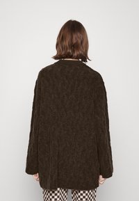 Braune, strukturierte Strickjacke mit lockerer Passform, die ein Zopfmuster und einen mittellangen Schnitt aufweist. Die Rückansicht zeigt überschnittene Schultern und lange Ärmel.