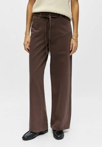 Pantalon large marron en coton, avec une taille à cordon, des poches avant et une texture lisse, porté avec des chaussures noires.