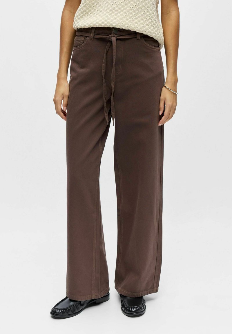 Pantalon large marron en coton, avec une taille à cordon, des poches avant et une texture lisse, porté avec des chaussures noires.