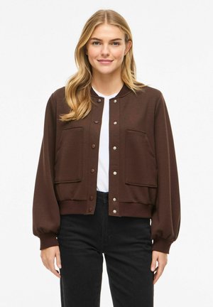 Kobieta o długich blond włosach, ubrana w brązową zapinaną na guziki kurtkę na białą koszulę i czarne spodnie, stojąca na tle gładkiego białego tła.