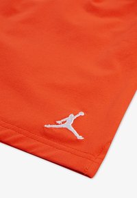 Oranžový sportovní dres s hladkým povrchem, na levém dolním rohu se nachází bílé vyšívané logo Jumpman.