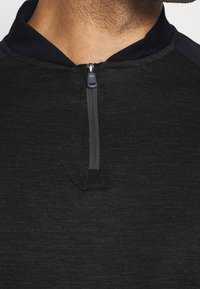 Musta urheilu paita, jossa on puoli-vetoketjukaulus, sileä pinta, tumman laivastonsinisiä yksityiskohtia olkapäillä ja vartalon myötäinen muoto.