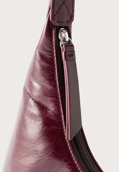 Gros plan sur un coin d'un sac en cuir bordeaux avec des coutures visibles, une fermeture éclair munie d'une languette en cuir et un fermoir en métal argenté.