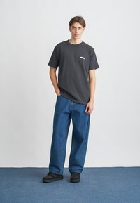 Μαύρο βαμβακερό μπλουζάκι με μικρό λευκό λογότυπο, συνδυασμένο με άνετο μπλε τζιν και μαύρα υποδήματα, ποζάροντας σε φόντο μπλε χαλιού.