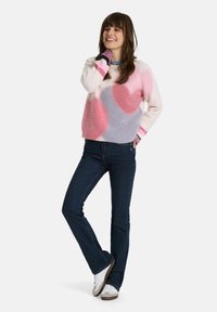 Flauschiger, mehrfarbiger Pullover mit großen pinken und lavendelfarbenen Formen, kombiniert mit dunkelblauen Jeans und weißen Sneakers. Strukturiertes Material und kontrastierende Bündchen.