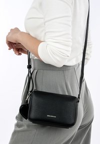 Schwarze Leder-Umhängetasche mit strukturiertem Äußeren, verstellbarem Schulterriemen und einem kleinen Silberemblem. Kompakte rechteckige Form.