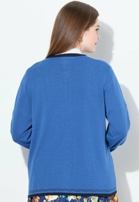 Blå stickad cardigan med enfärgad design, långa ärmar, rund halsringning och dekorativ detalj vid fållen.