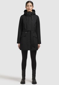 Schwarzer wasserdichter Parka mit verstellbarer Kapuze, tailliertem Gürtel, zwei Fronttaschen und Druckknopfverschluss, kombiniert mit schwarzen Leggings und Stiefeln.