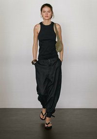Black sleeveless top paired with loose black trousers and flip-flops. The outfit is complemented by a green shoulder bag and wooden bangles.