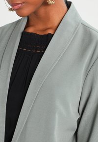 Cardigan grigio chiaro con una texture liscia e design con apertura frontale, abbinato a un top nero con un colletto in crochet dettagliato.