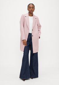 Γυναίκα που στέκεται φορώντας ένα ανοιχτό ροζ παλτό μήκους μέχρι το γόνατο, λευκό τοπ, σκούρο τζιν με φαρδιά παντελόνια και σκουρόχρωμα μυτερά παπούτσια σε απλό λευκό φόντο.