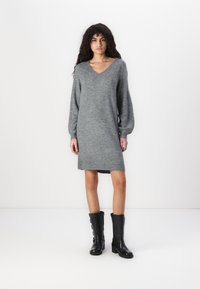 Robe en tricot grise avec un décolleté en V, des manches longues et un ourlet côtelé, associée à des bottes en cuir noires jusqu'au genou avec des zips latéraux.