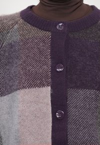 Paars geruite cardigan met een ronde halslijn, gestructureerde stof en drie paarse knopen aan de voorkant. Zacht, wolachtig materiaal.