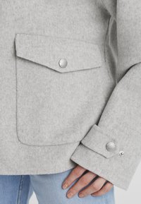 Graue gefütterte Jacke mit zwei Fronttaschen, silbernen Druckknöpfen und einem lockeren Schnitt. Sie hat eine weiche Textur und umgeschlagene Ärmel.