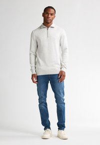 Pullover grigio chiaro con colletto a metà zip, polsini e orlo a costine, abbinato a jeans in denim blu e sneakers bianche. Texture liscia.