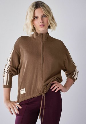 Frau mit kurzem blondem Haar, die einen braunen Reißverschluss-Pullover mit weißen Streifen an den Ärmeln und lila Hose trägt, posiert mit einer Hand in der Hüfte.