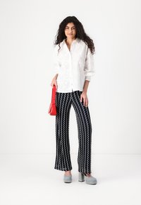 White embroidered shirt with long sleeves, paired with navy patterned pants. Model holds a red handbag and wears gray platform heels.
