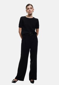 Sort, kortærmet striktrøje sat sammen med højtaljede, vide sorte bukser og spidse sorte flade sko. Bælte tilføjer struktur.