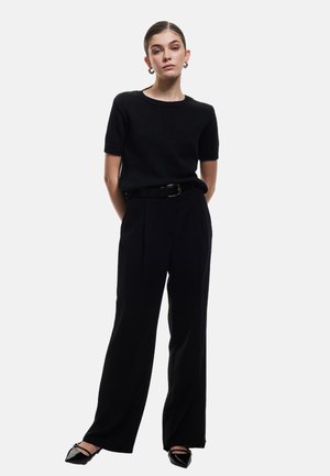 Top in maglia nera a maniche corte abbinato a pantaloni neri a vita alta e a gamba larga, insieme a ballerine nere a punta. La cintura aggiunge struttura.